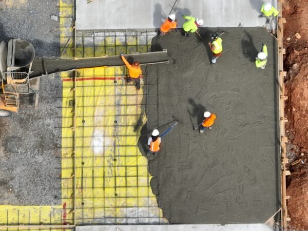 Construction workers pour and spread fresh concrete on a foundation, with a concrete truck and reinforcement grid visible.